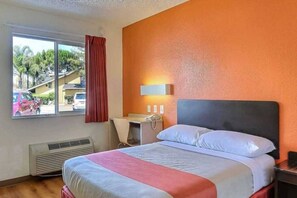 A hotel room with a bed, a desk, a chair, a window with a view of the outside, and a wall-mounted lamp.
