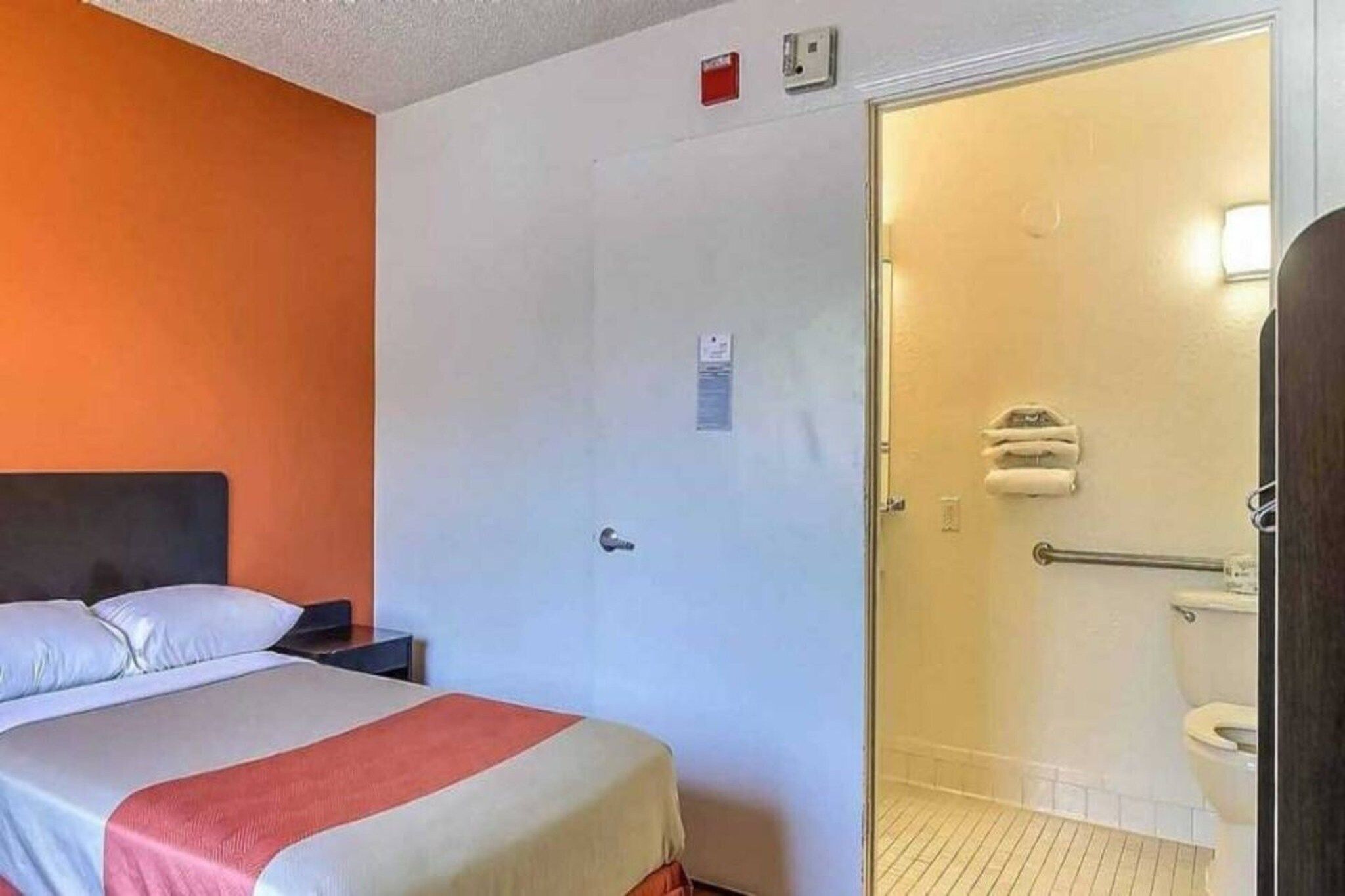 Quarto de hotel com cama, mesa de cabeceira e porta para banheiro com vaso sanitário e chuveiro.