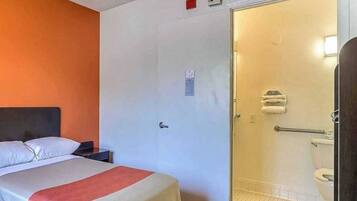 Quarto de hotel com cama, mesa de cabeceira e porta para banheiro com vaso sanitário e chuveiro.