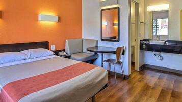A hotel room with a bed, a desk, a chair, a small table, and a bathroom with a sink.