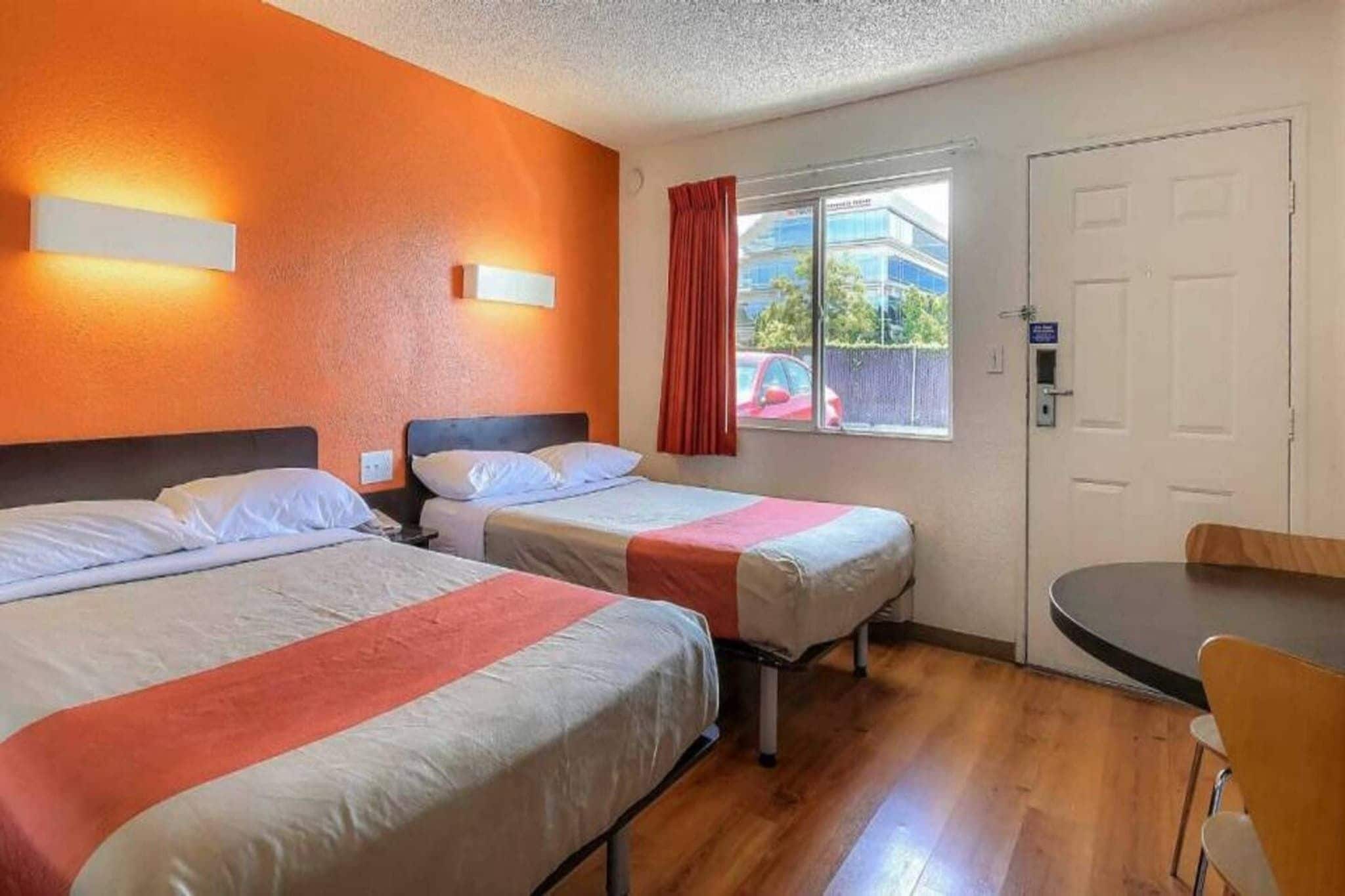 A hotel room with two beds, a window with a view of the outside, a door, and a small table with a chair.