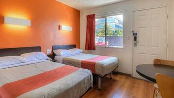 A hotel room with two beds, a window with a view of the outside, a door, and a small table with a chair.