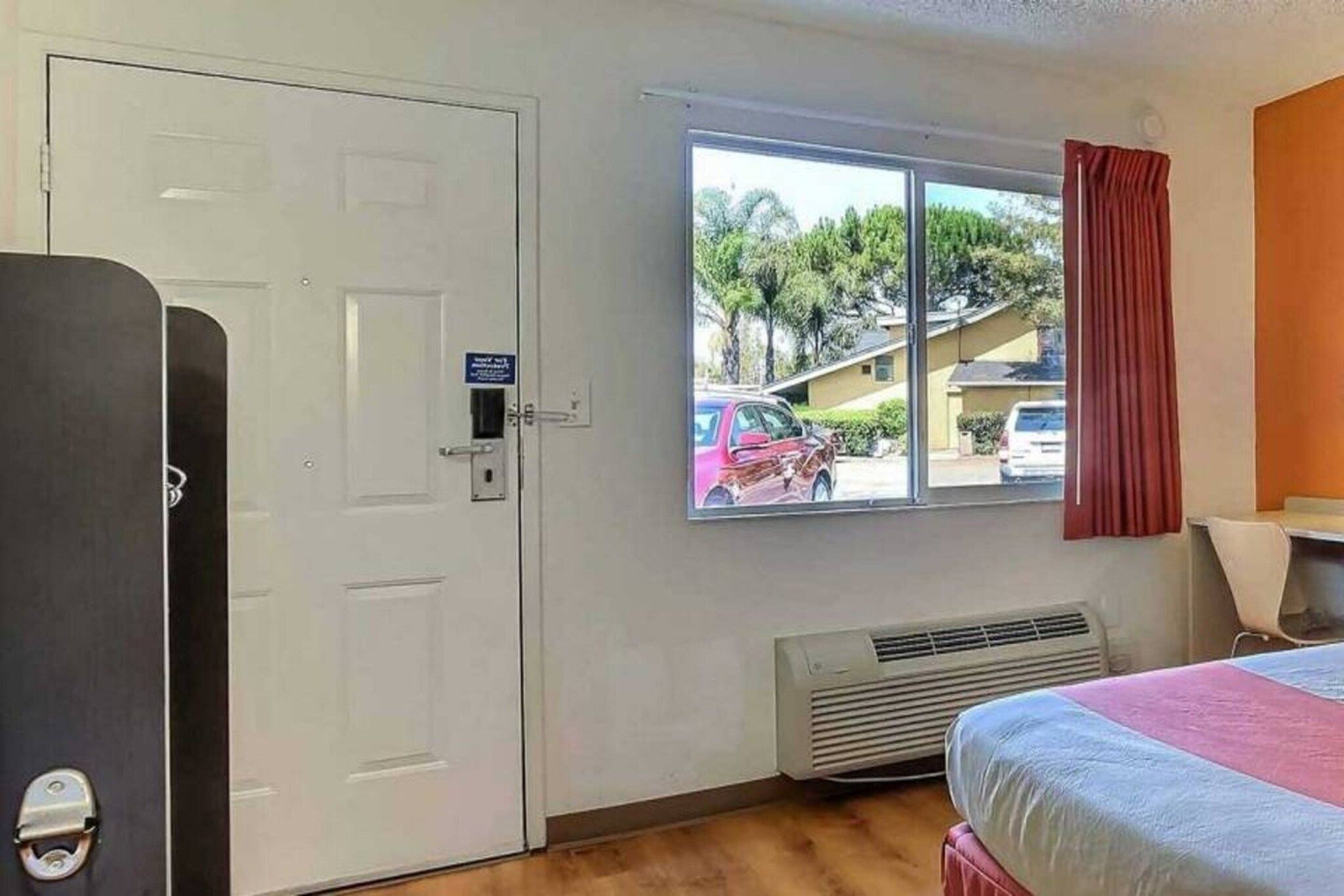A room with a bed, a window with a view of parked cars and houses, a white door, and a heater.