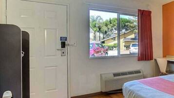 A room with a bed, a window with a view of parked cars and houses, a white door, and a heater.