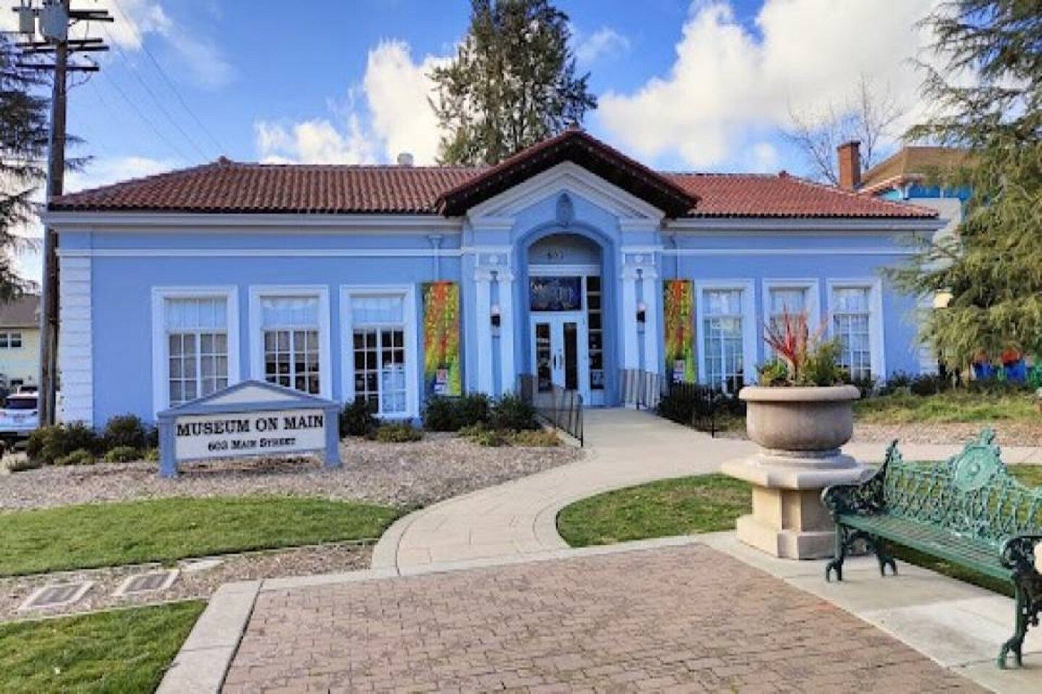 A building with a blue facade, a sign reading 'Museum on Main', and a paved entrance area with a bench.