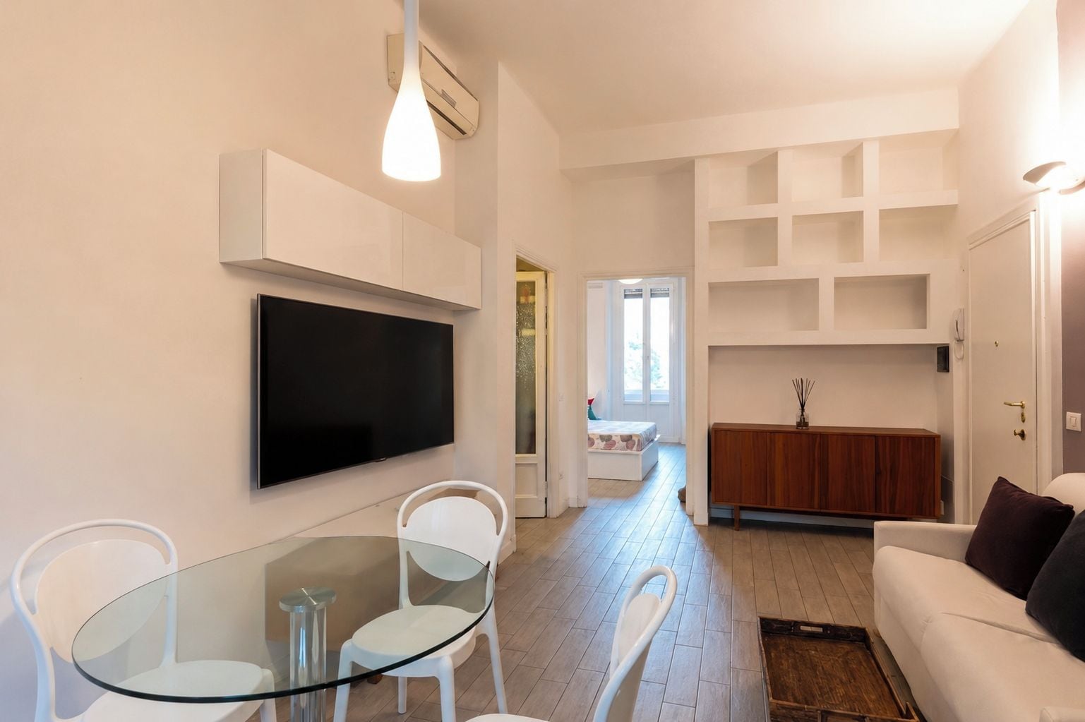 A modern living room with a glass dining table, white chairs, a flat-screen TV mounted on the wall, and a wooden cabinet.