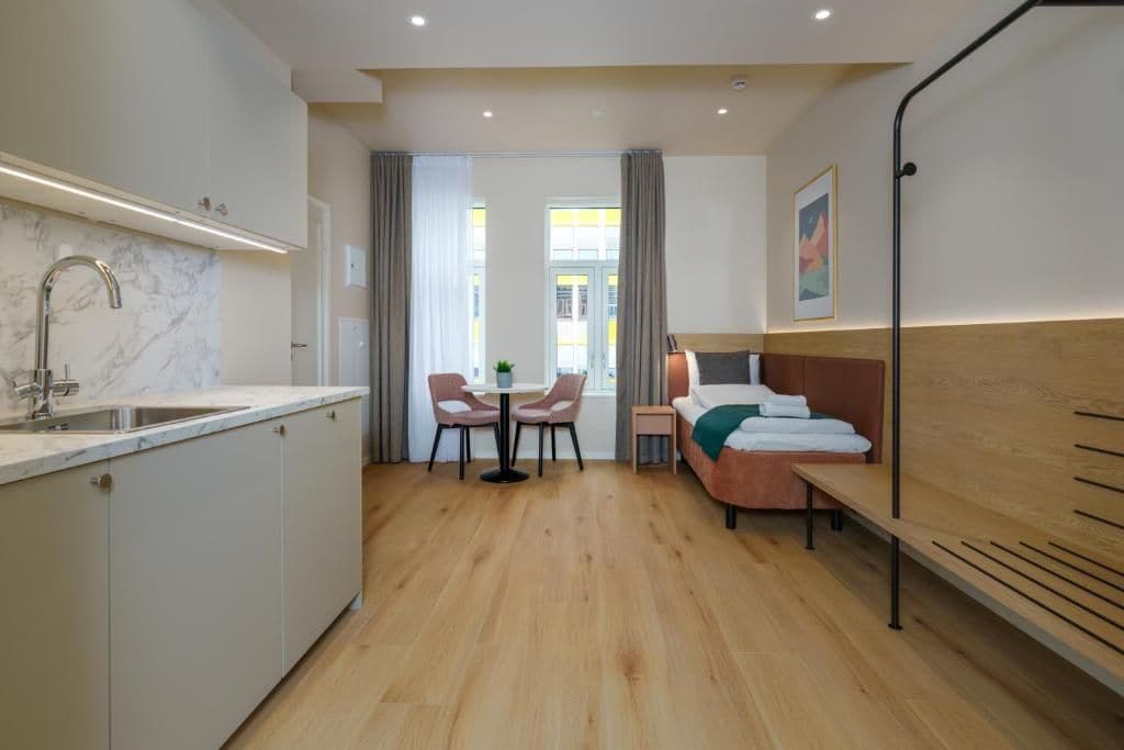 Habitació d'hotel compacta amb cuina, taula de menjador i cadires, llit amb capçalera i finestra amb cortines.