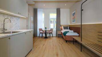 Habitació d'hotel compacta amb cuina, taula de menjador i cadires, llit amb capçalera i finestra amb cortines.
