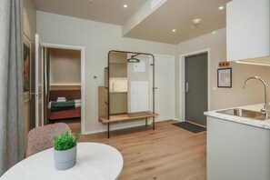 Una habitación de hotel moderna con suelo de madera, una mesa de comedor redonda, una pequeña planta en maceta, una cama con manta verde, un espejo, una silla colgante, un banco empotrado, un lavamanos y una puerta que da a un baño.