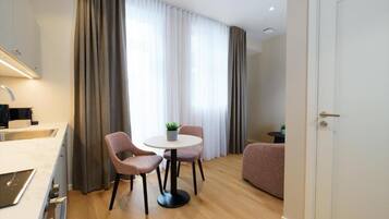 Habitación de hotel moderna con kitchenette, zona de comedor con mesa y sillas, sofá y un ventanal con cortinas.