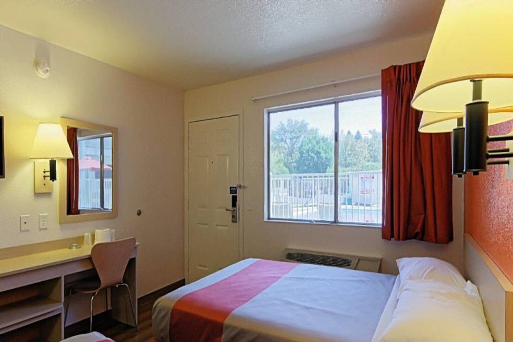 A hotel room with a bed, a desk, a chair, a window with curtains, and a lamp.