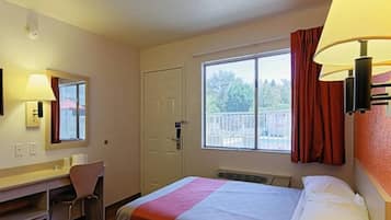 A hotel room with a bed, a desk, a chair, a window with curtains, and a lamp.