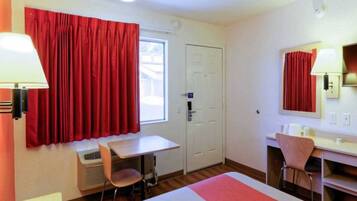 Une chambre d’hôtel avec un lit, un bureau, une chaise, une fenêtre avec des rideaux rouges et une porte.