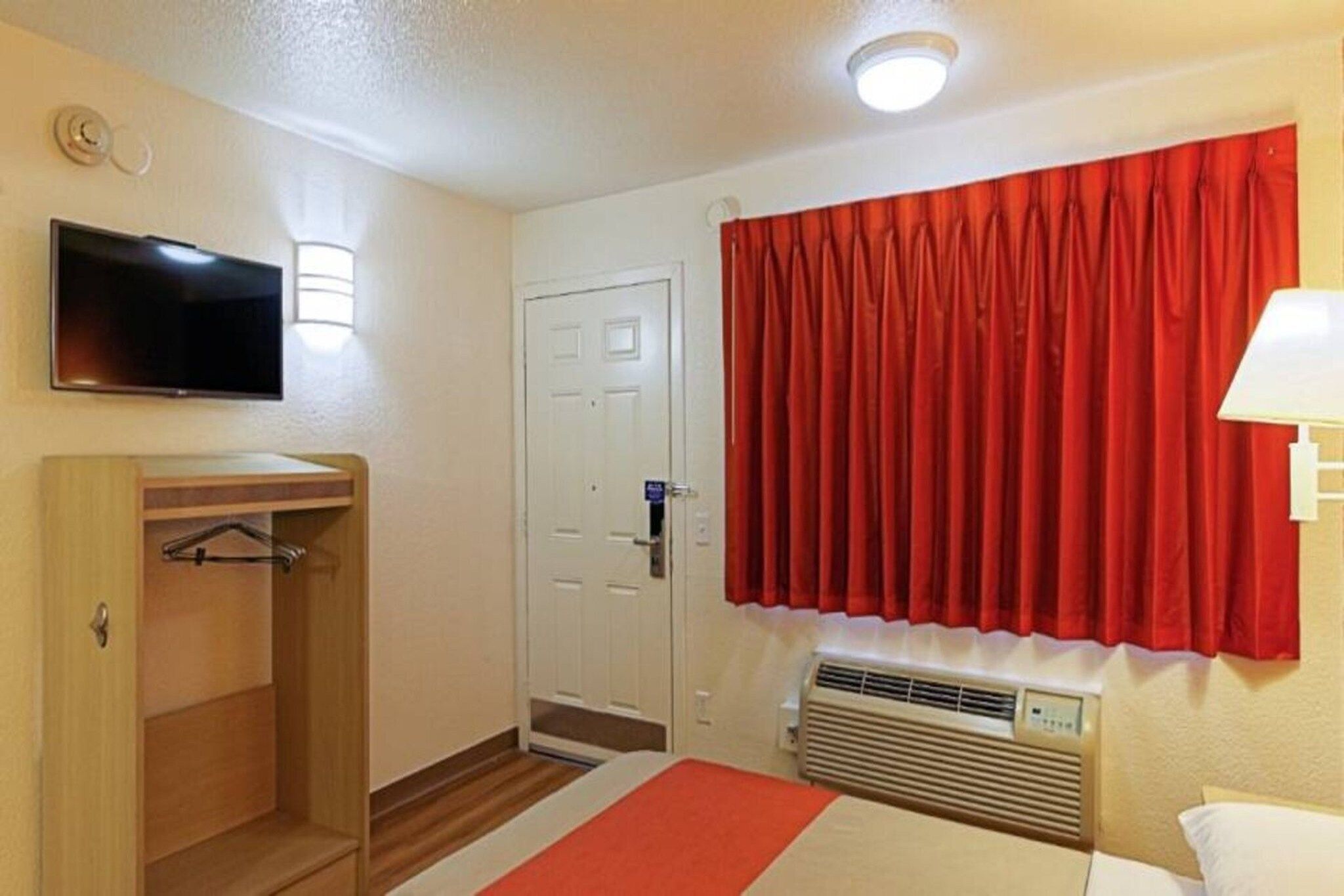 A hotel room with a bed, a television, a wardrobe, a door, a window with red curtains, and a wall-mounted air conditioning unit.