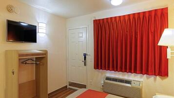 A hotel room with a bed, a television, a wardrobe, a door, a window with red curtains, and a wall-mounted air conditioning unit.
