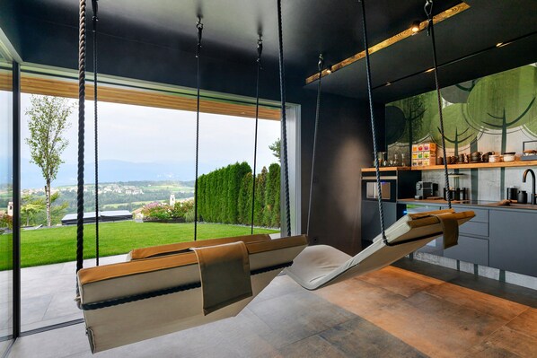 A modern kitchen with a hanging chair and a large window overlooking a green landscape.