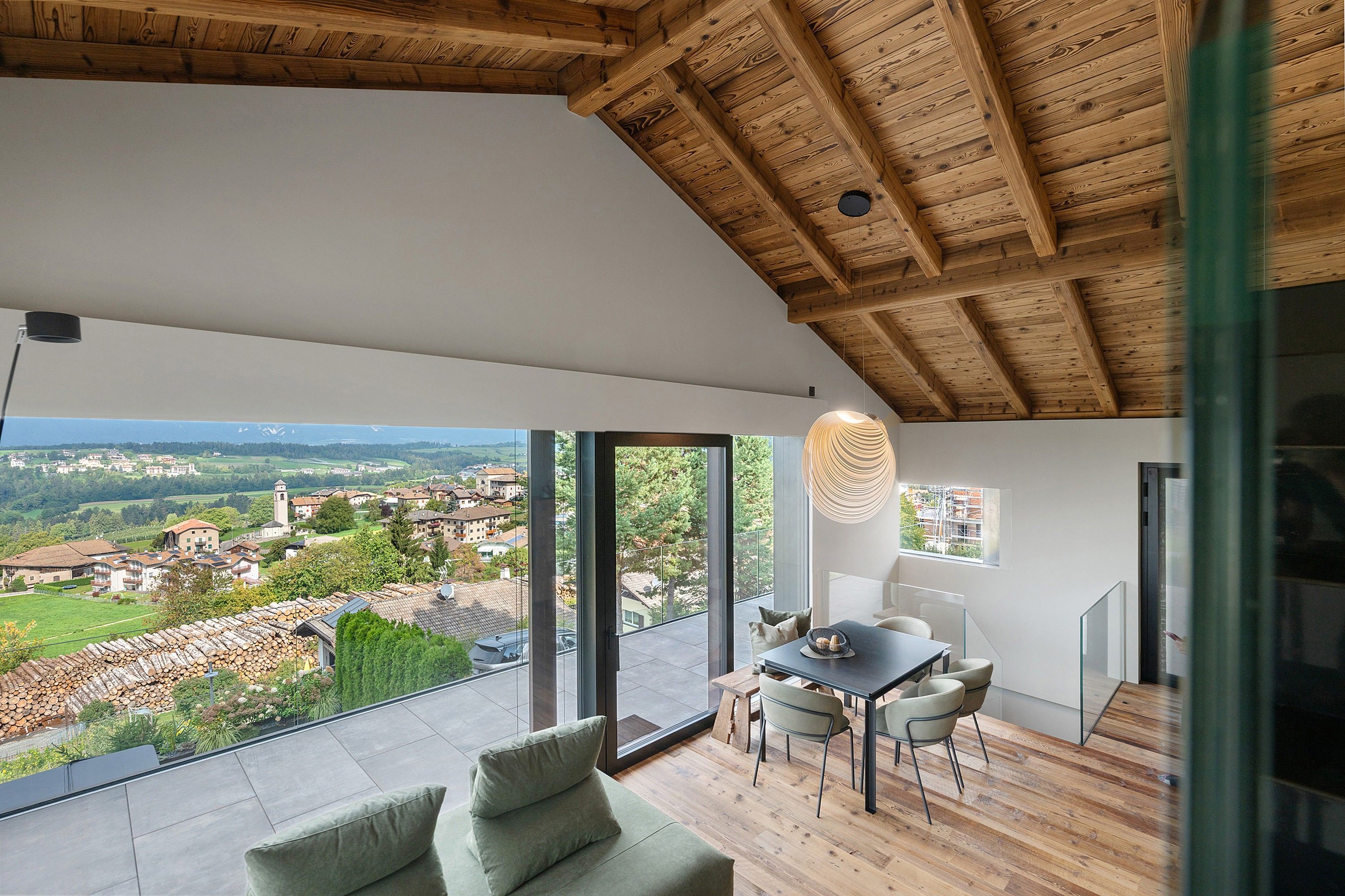 A modern interior with a wooden ceiling, a dining area with a table and chairs, and a large window offering a view of a village.