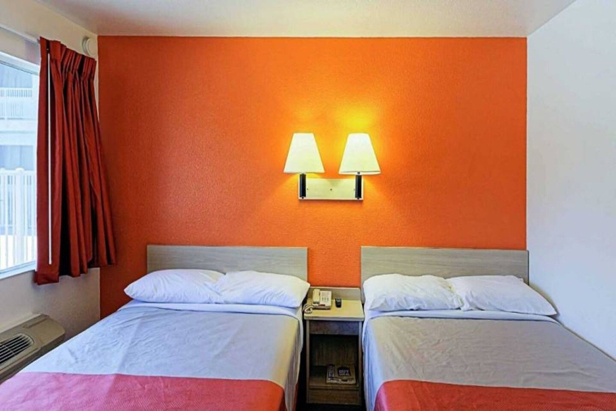 Habitación de hotel con dos camas, una lámpara de pared y un ventanal con cortinas.