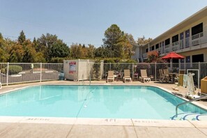 Ein rechteckiger Swimmingpool mit einer Tiefenmarkierung von etwa 1,2 Metern, umgeben von einem weißen Zaun, und ein Gebäude mit Balkonen im Hintergrund.
