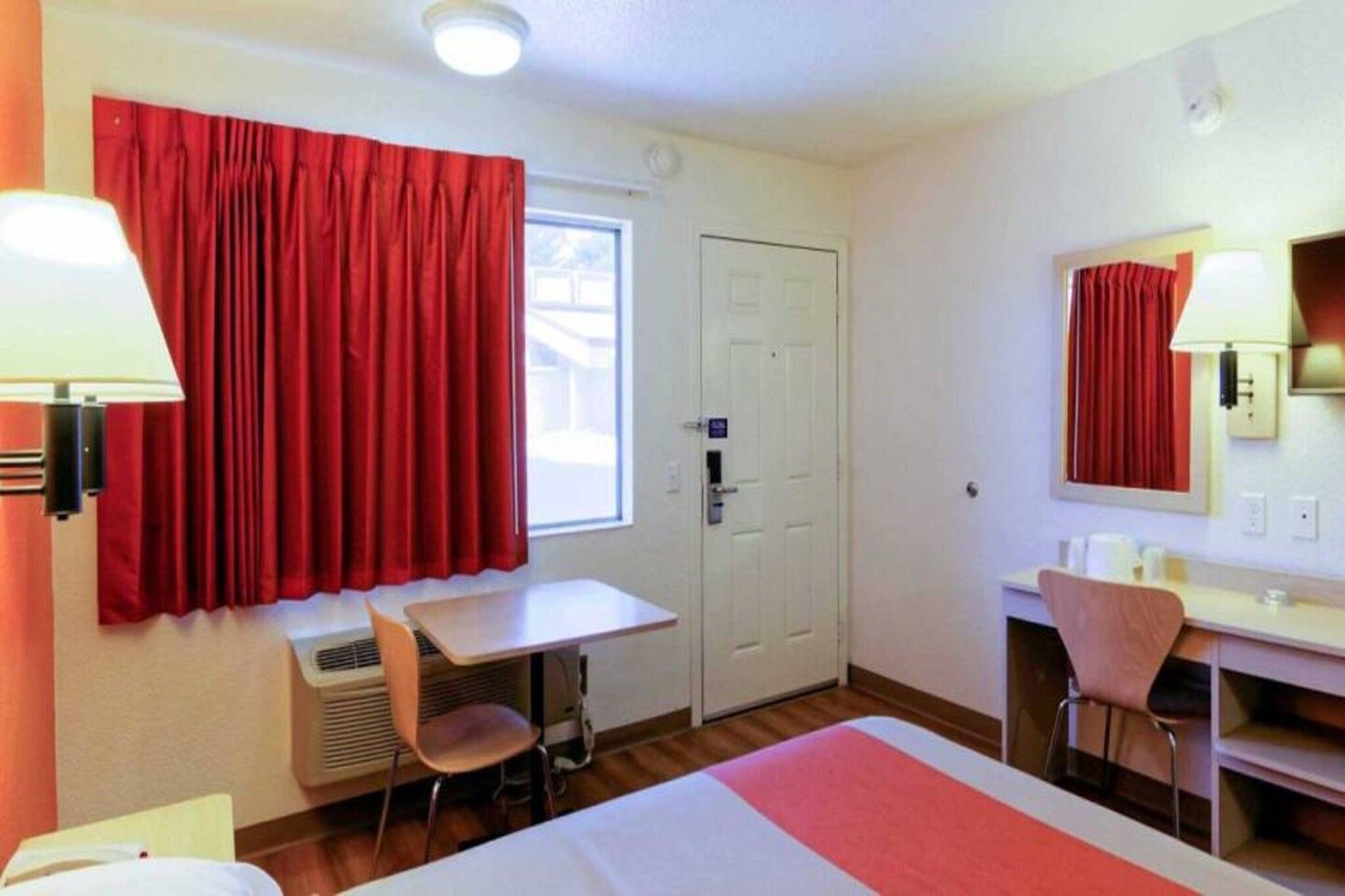 Une chambre d’hôtel avec un lit, un bureau, une chaise, une fenêtre avec des rideaux rouges et une porte.