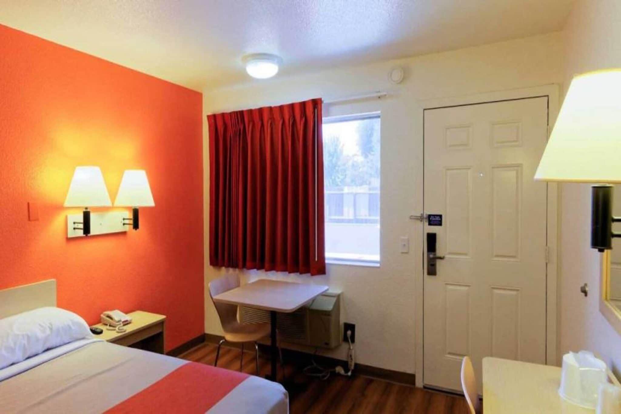 Habitación de hotel con cama, mesita de noche, lámpara, escritorio, silla y ventana con cortinas rojas.