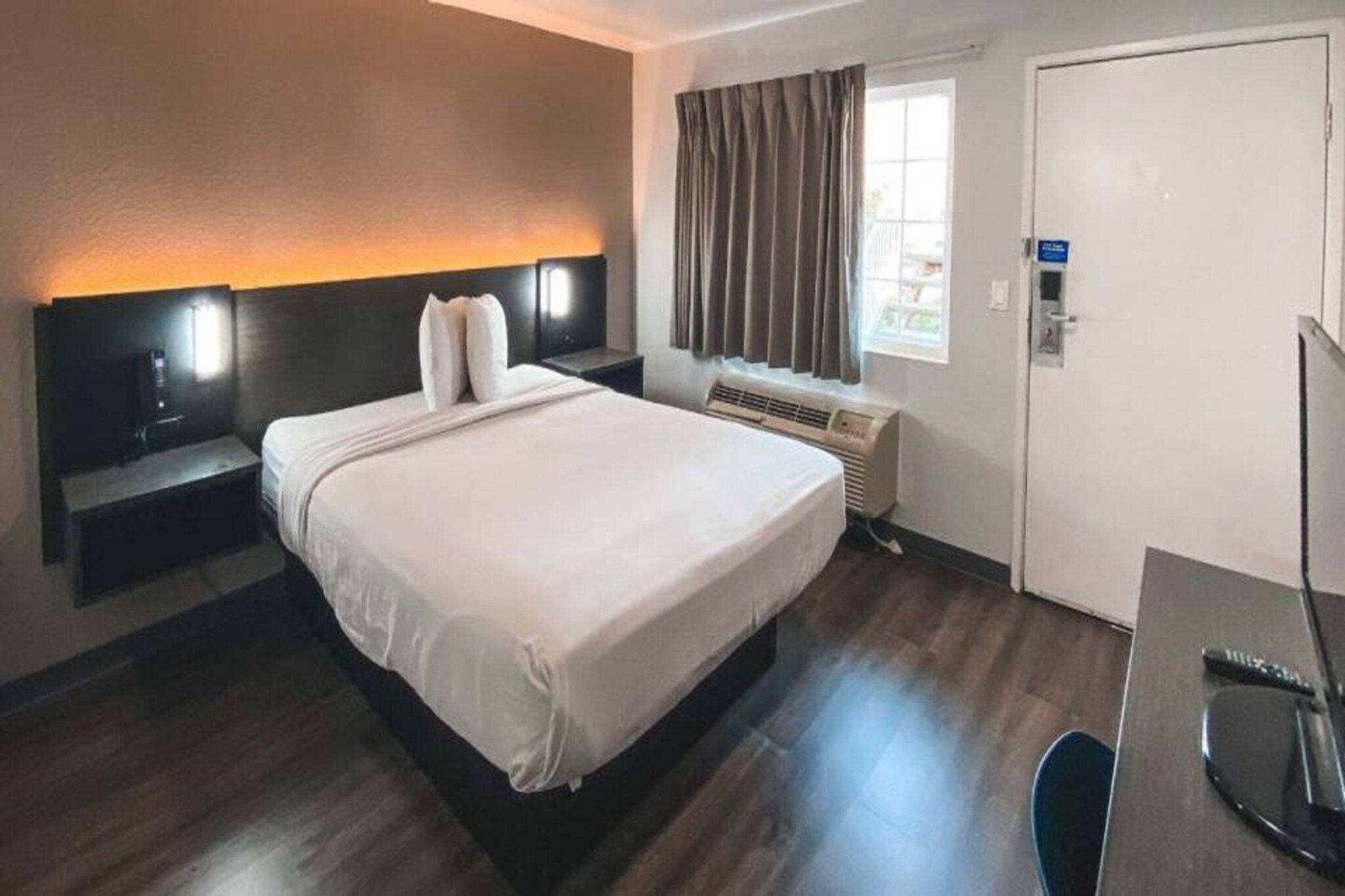 A hotel room with a bed, bedside table, lamp, window with curtains, and a door.