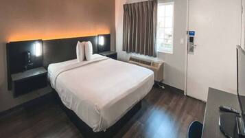 A hotel room with a bed, bedside table, lamp, window with curtains, and a door.