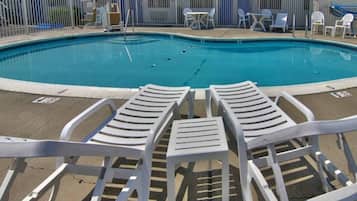 Piscina com cadeiras de descanso e mesas, cercada por uma grade.