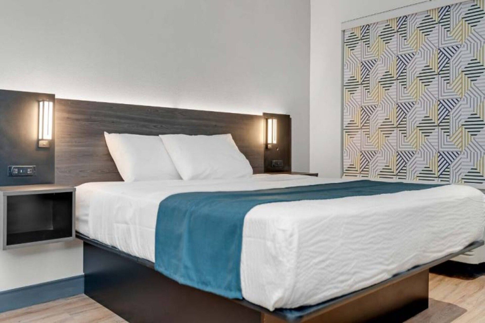 A modern hotel room with a large bed, a nightstand, a wall-mounted light, and a patterned wall.
