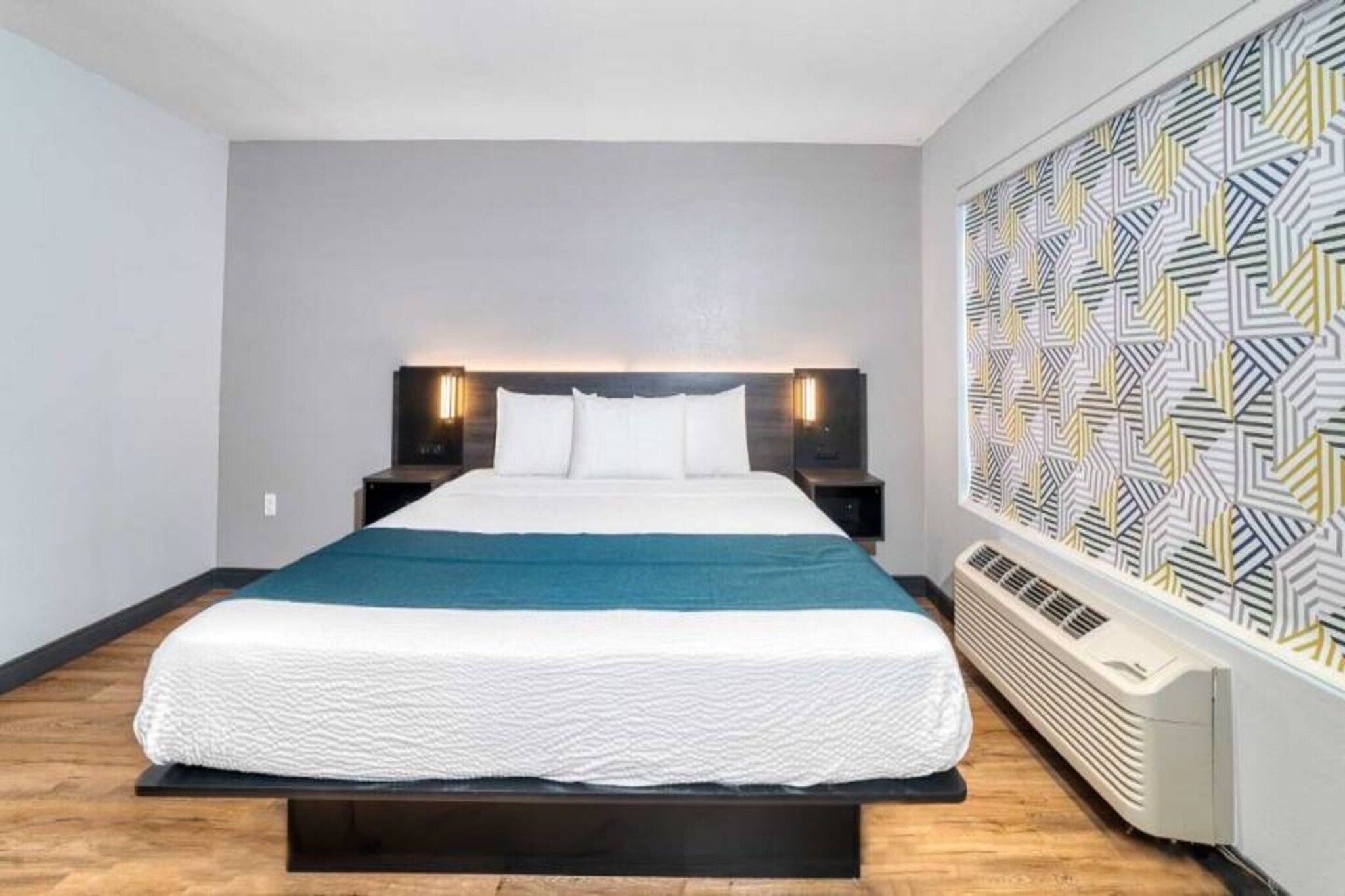 Ein modernes Hotelzimmer mit einem großen Bett, einer Kopfteilbeleuchtung, einer Wand mit Muster und einer fest installierten Klimaanlage.