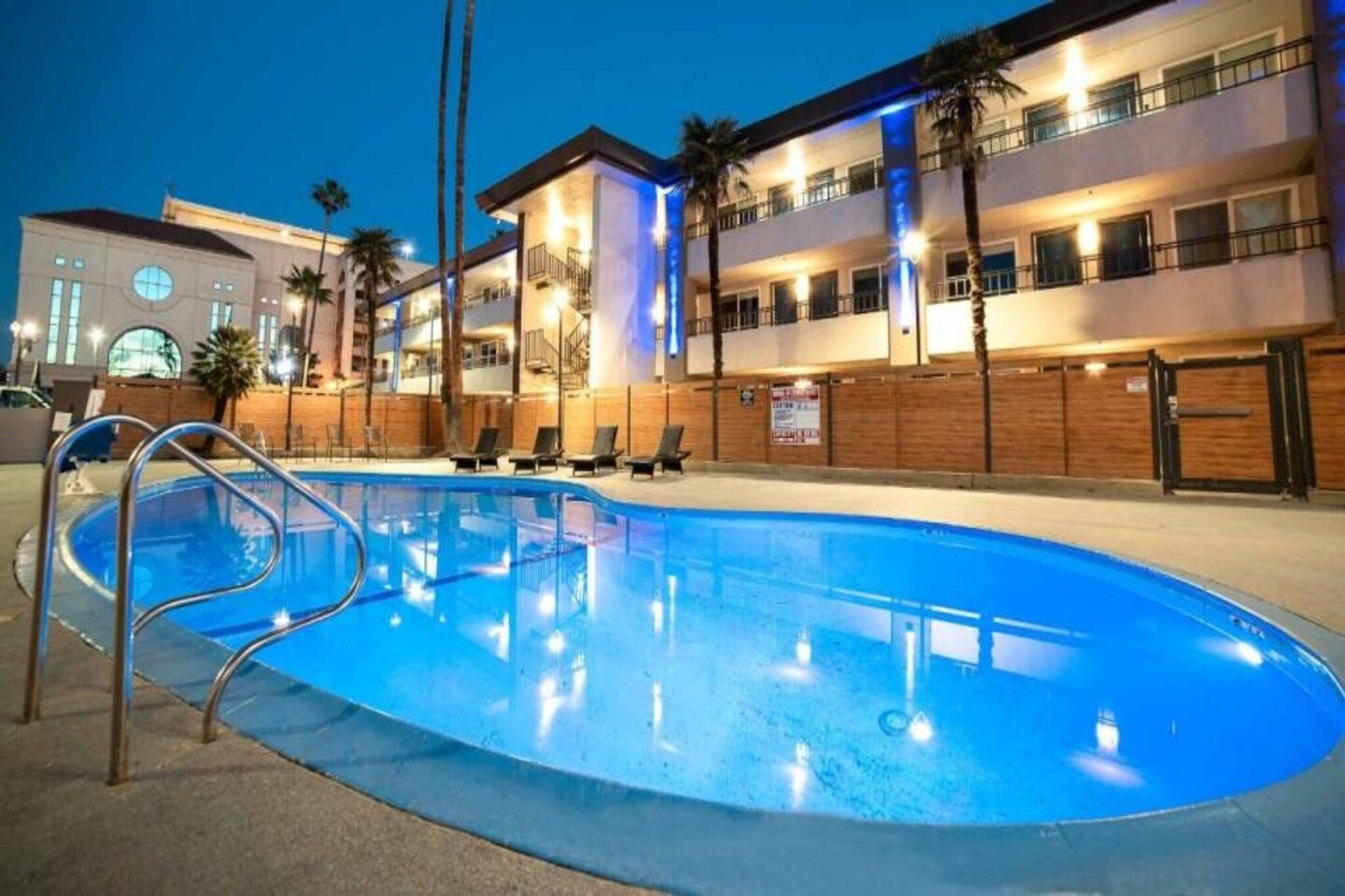 Una piscina con una escalera metálica, rodeada de sillas de descanso y palmeras, con un edificio de varios pisos al fondo.