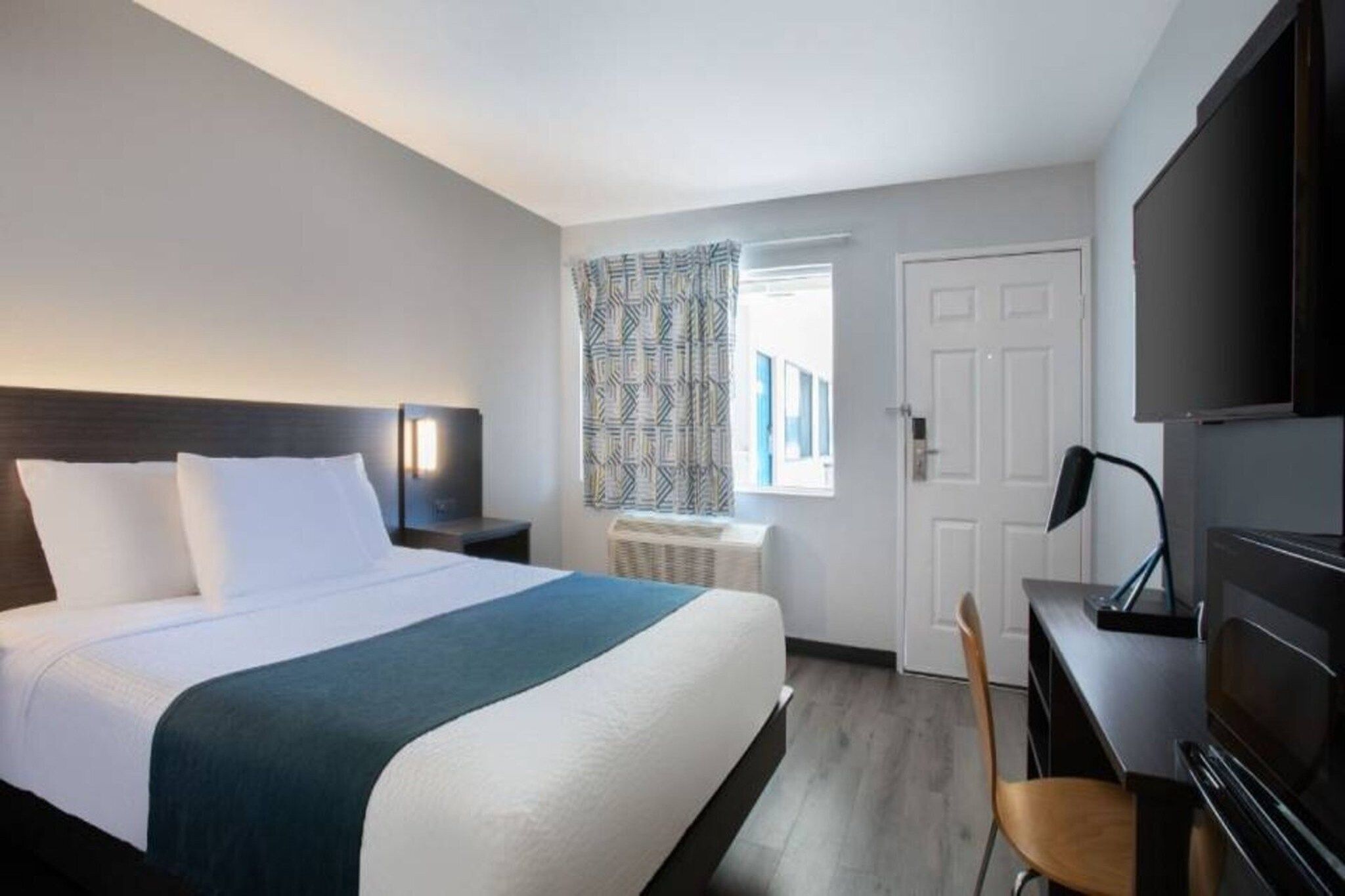Una habitación de hotel con cama, escritorio, silla, televisor y ventana con cortinas.