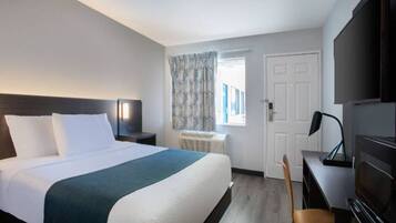 Una habitación de hotel con cama, escritorio, silla, televisor y ventana con cortinas.