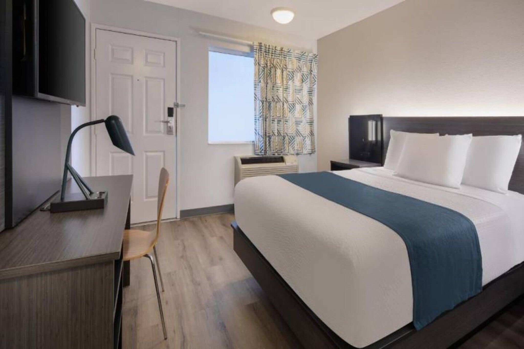 A hotel room with a bed, a desk, a chair, a TV, and a window with patterned curtains.