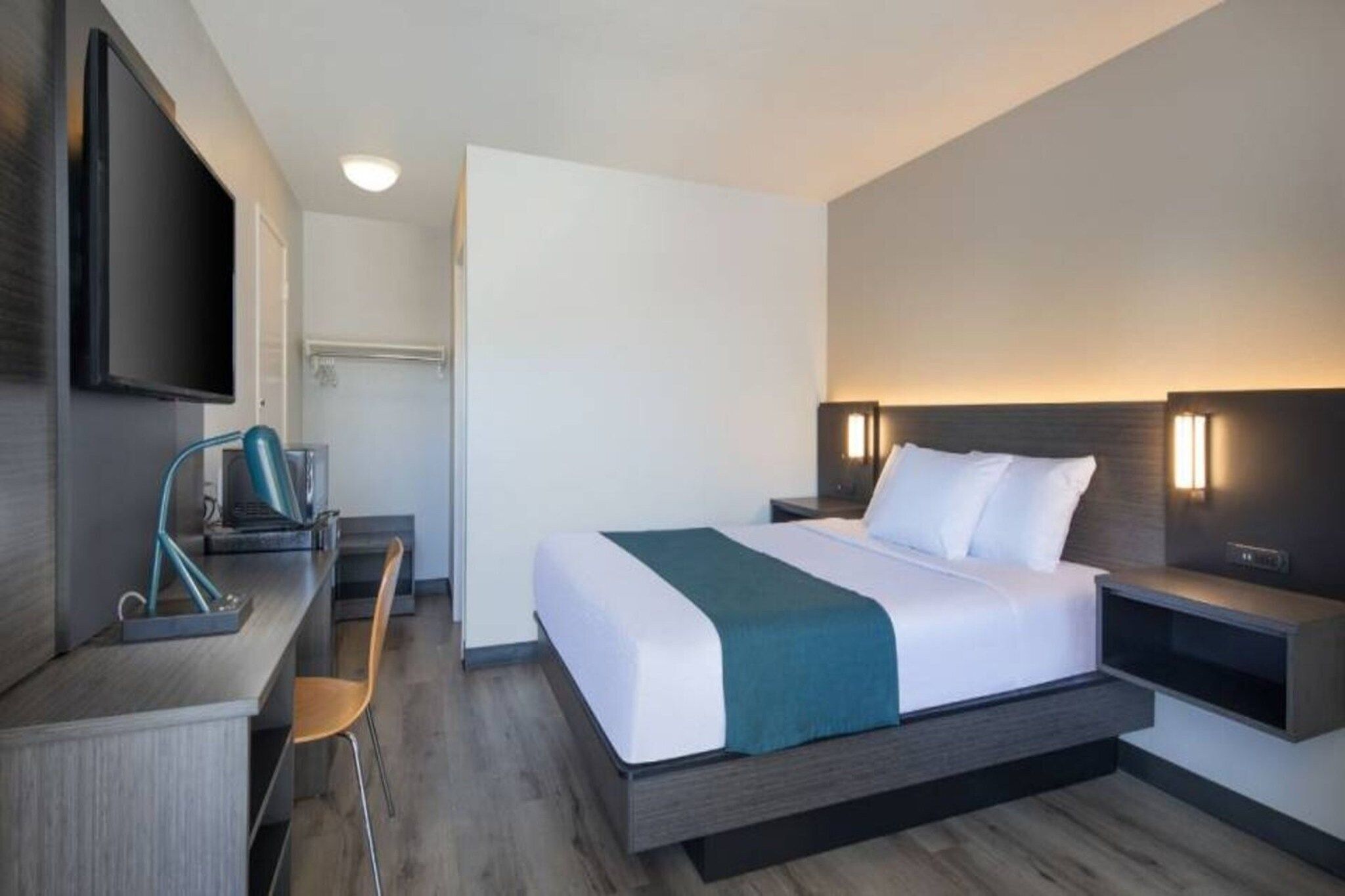 A hotel room with a bed, bedside tables, a desk with a chair, a television, and a wall-mounted light fixture.