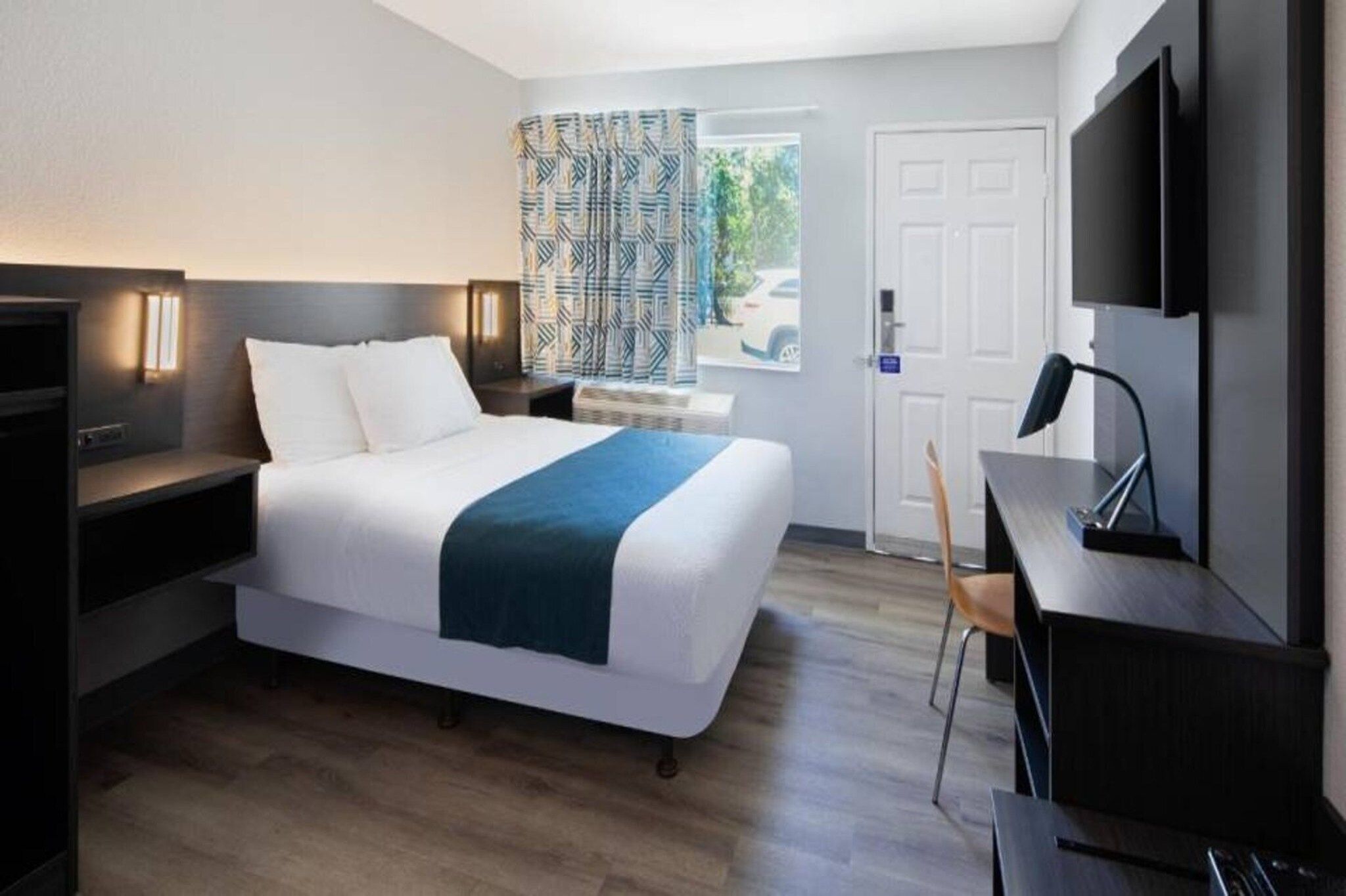 A hotel room with a bed, bedside tables, a desk, a chair, a TV, and a door.
