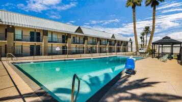 A hotel with a swimming pool, palm trees, and a covered seating area.