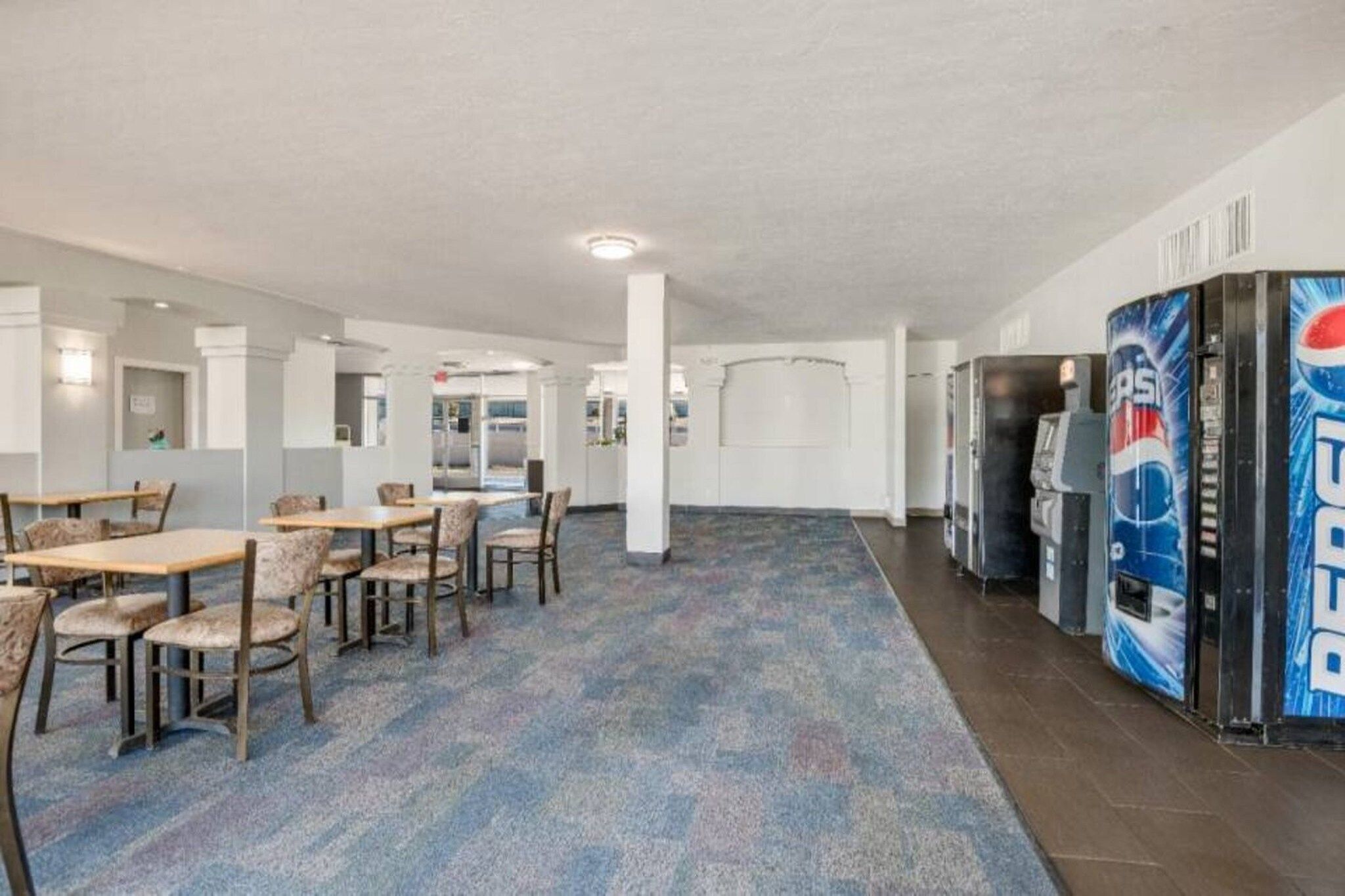 A hotel lobby with a carpeted floor, seating area with tables and chairs, vending machines, and a large pillar.