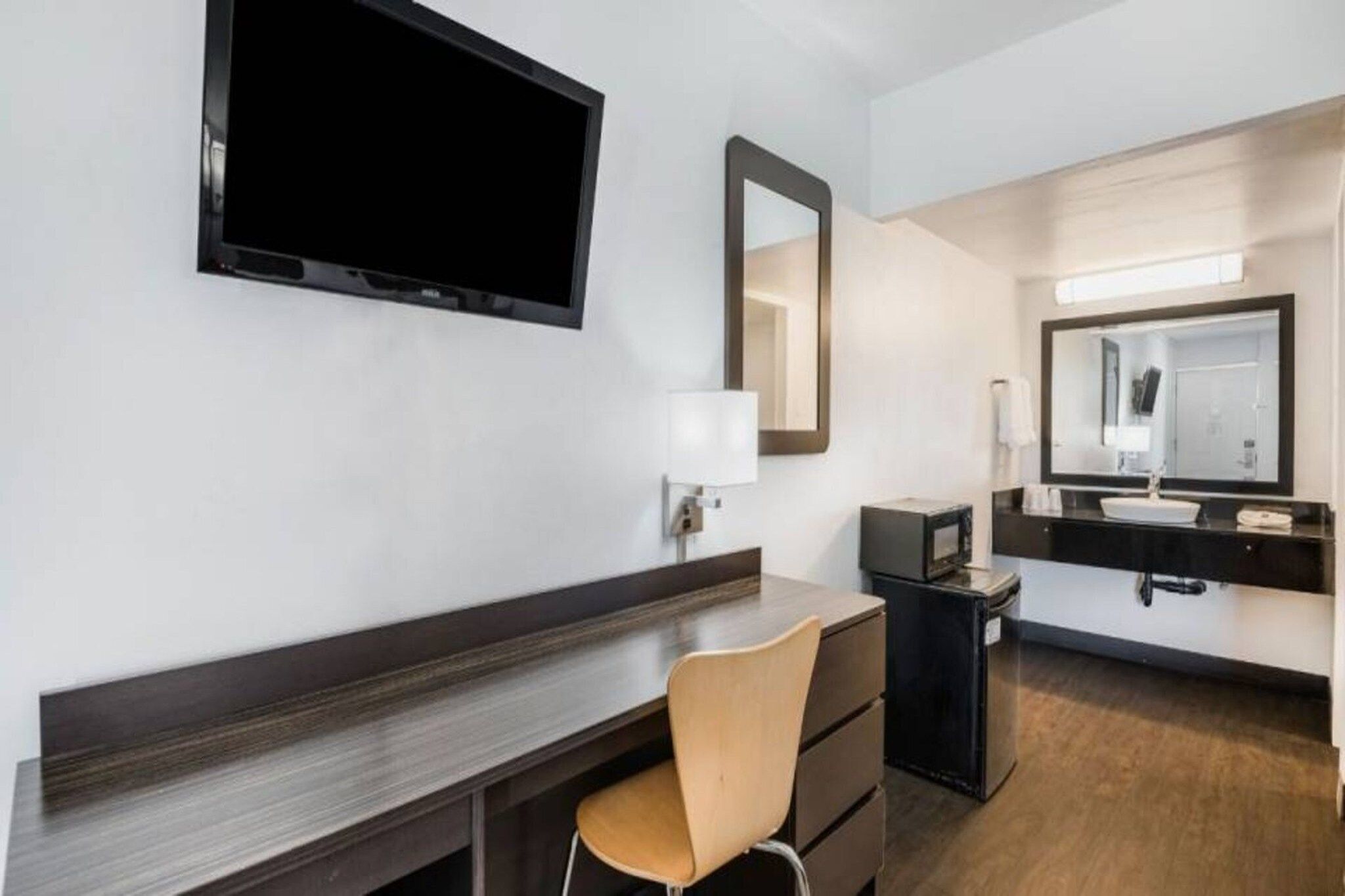 A hotel room with a flat-screen TV mounted on the wall, a desk with a chair, a dresser, a vanity with a mirror, and a small refrigerator.