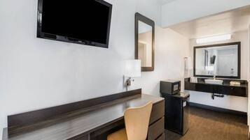 A hotel room with a flat-screen TV mounted on the wall, a desk with a chair, a dresser, a vanity with a mirror, and a small refrigerator.