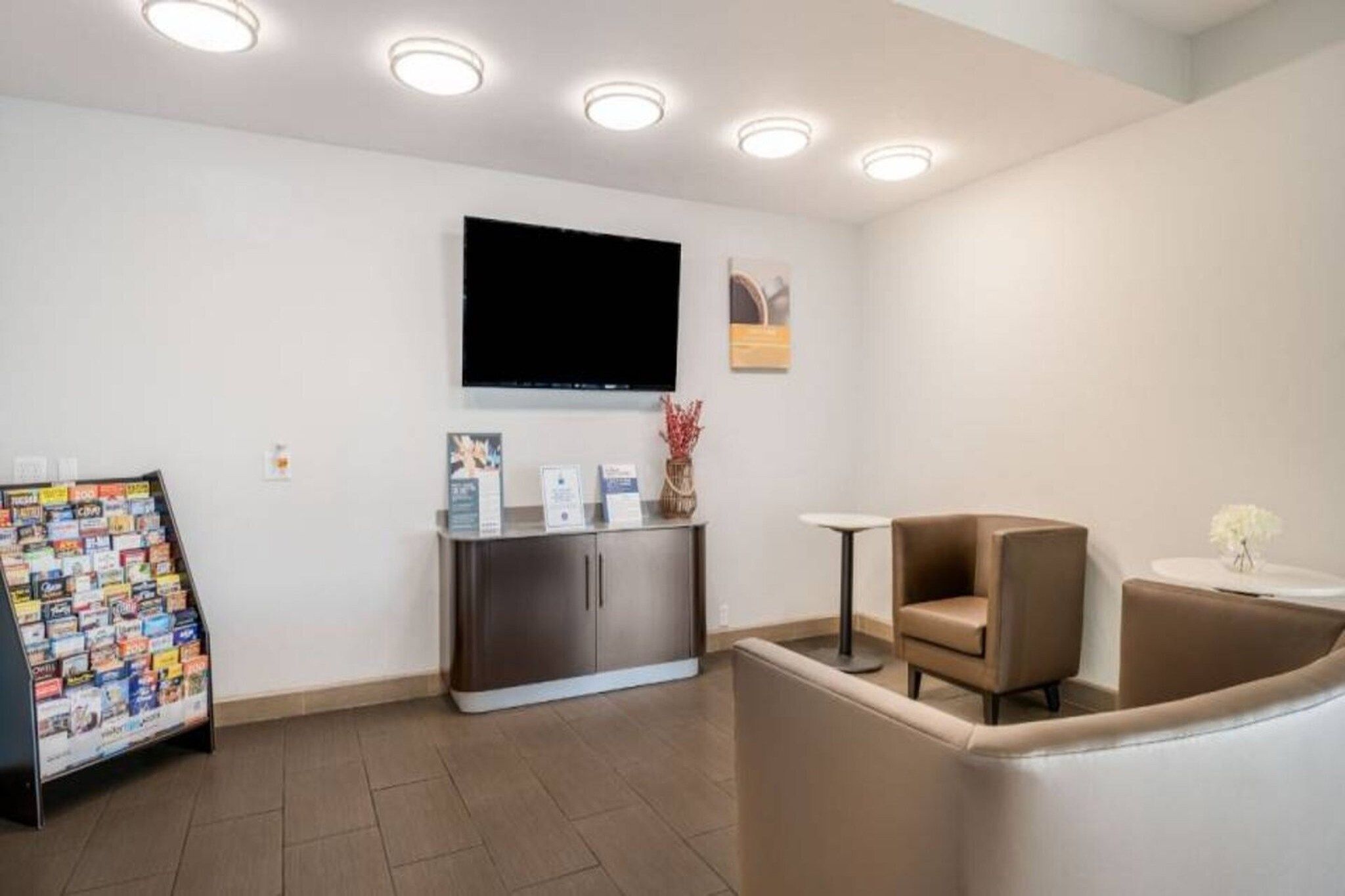 A reception area with a flat-screen TV mounted on the wall, a magazine rack, a small table with a vase, and a cushioned seating area.