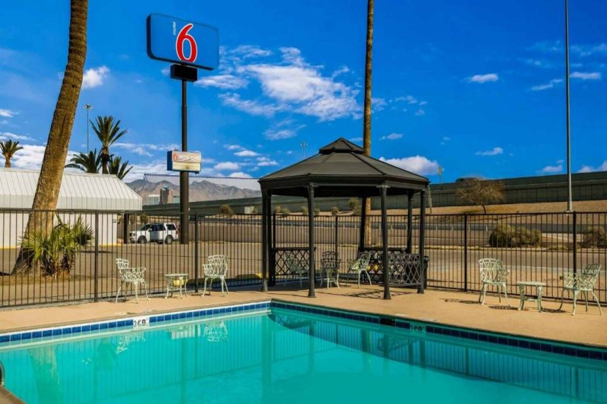 A clear blue pool with lounge chairs and a gazebo, surrounded by a fenced area with palm trees and a mountain in the background.