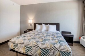 A hotel room with a bed, a bedside table with a lamp, and a heater. - 1 King Bed | Non-Smoking, Mini Fridge (Tucson AZ)