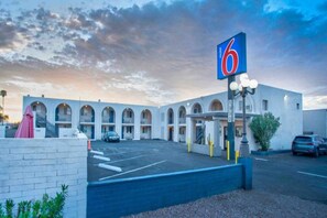 Un motel avec une rangée de fenêtres en arc de cercle, un parking et un panneau indiquant le numéro 6.