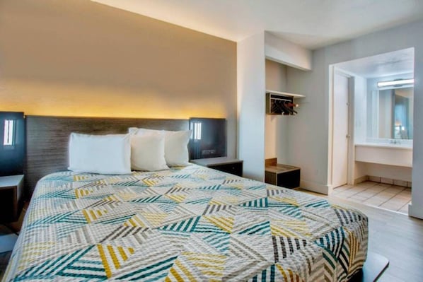 Un dormitorio con una cama grande, una mesita de noche, un estante en la pared y una puerta que da a otra habitación.