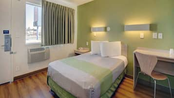 A hotel room with a bed, a desk, a chair, a window with curtains, and a wall-mounted heater.