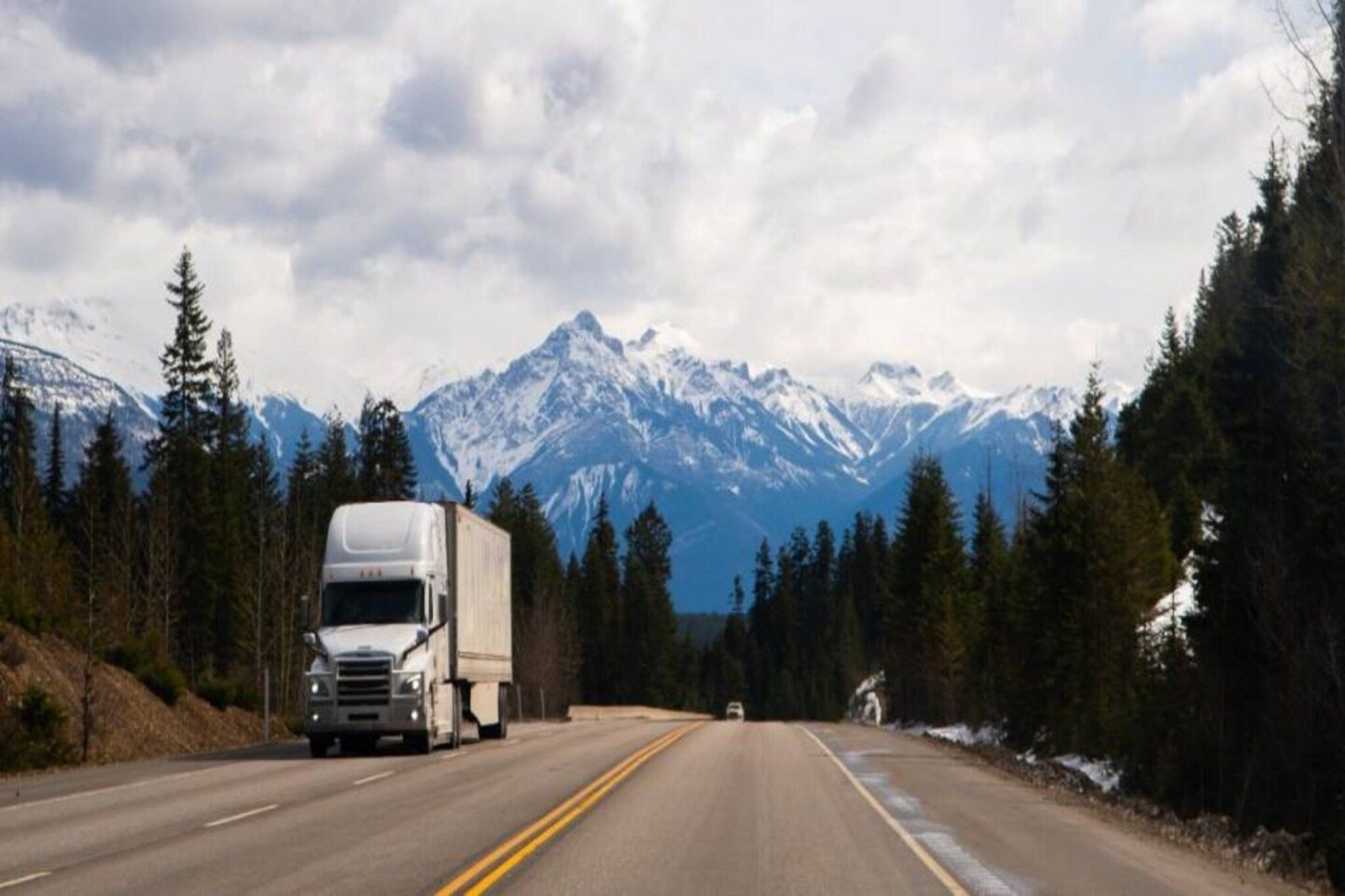 A road with a large truck and a smaller vehicle, surrounded by snow-capped mountains and dense forests.