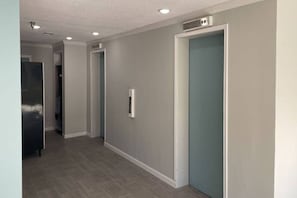 Een gang met een grijze muur, een deur met een blauwe hendel en een roestvrijstalen apparaat aan de linkerkant.