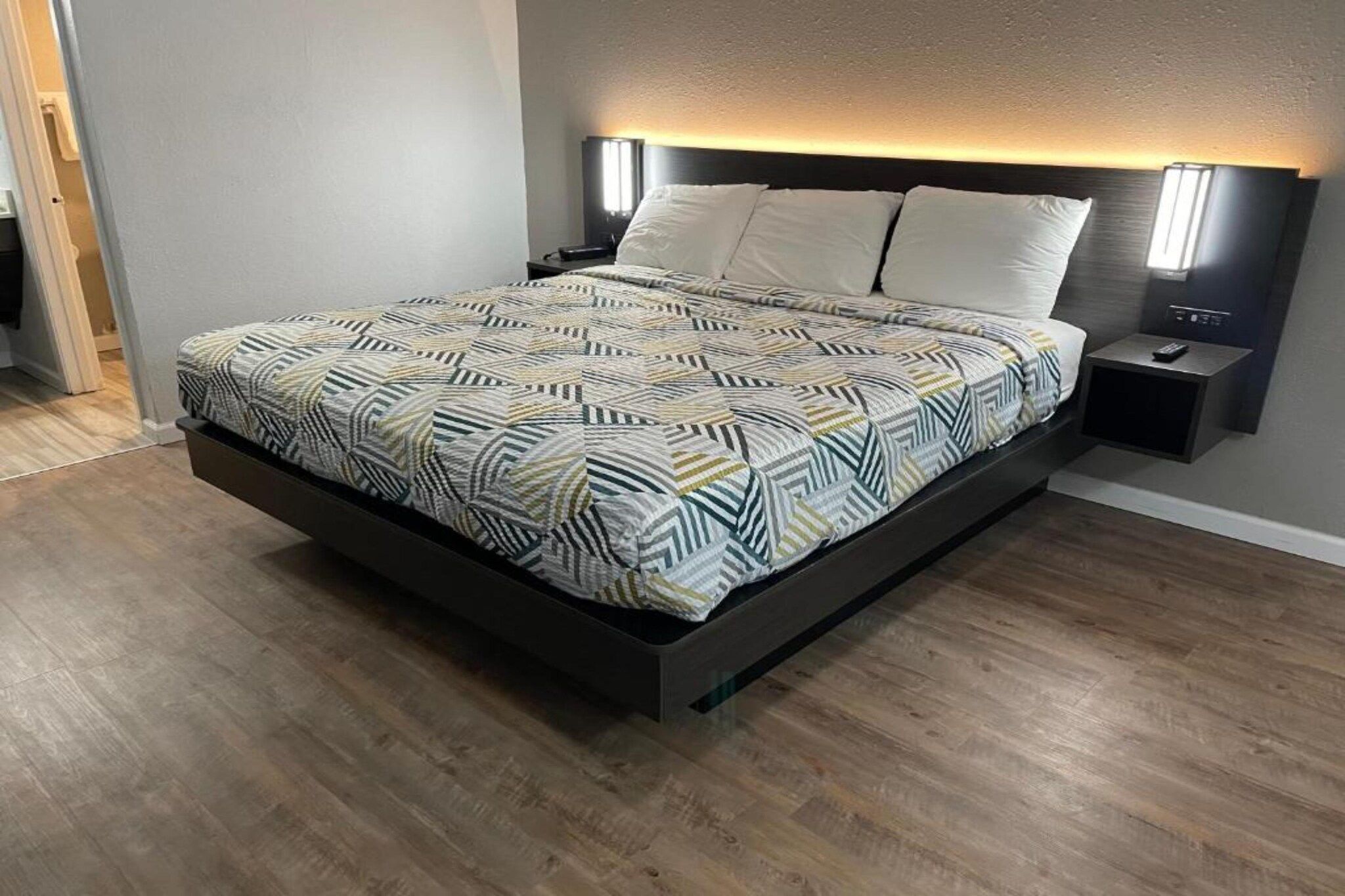 A bed with a patterned bedspread, a bedside table with a lamp, and a nightstand with a remote control.