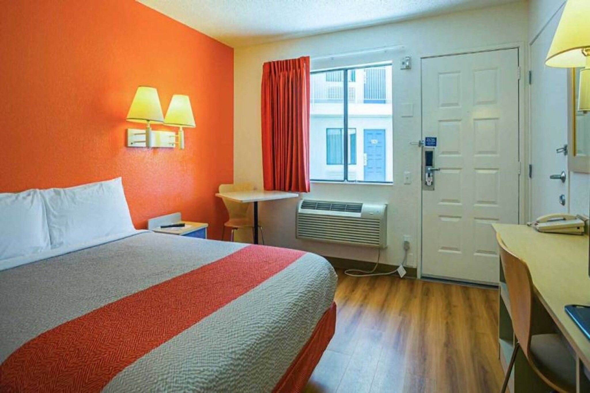 A hotel room with a bed, a desk, a chair, a window with curtains, and a door.